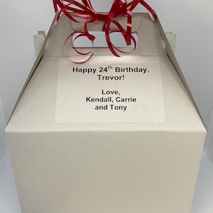 Puede incluir: Una caja de regalo blanca con una cinta roja en la parte superior. La caja tiene una etiqueta que dice "Happy 24th Birthday, Trevor! Love, Kendall, Carrie and Tony."