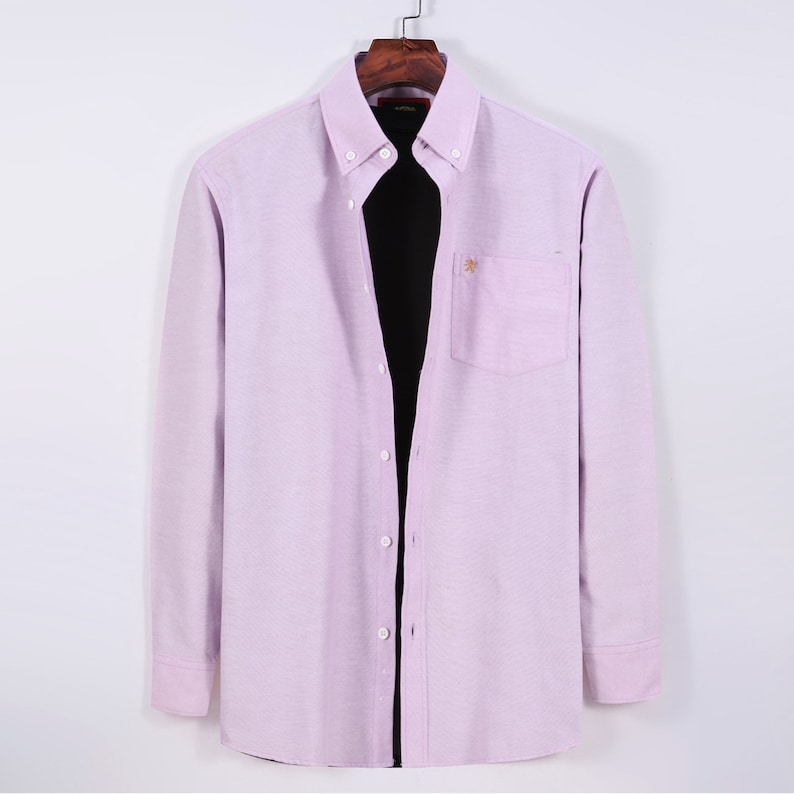 Puede incluir: Camisa abotonada de color lila claro con mangas largas, bolsillo en el pecho y un peque&ntilde;o detalle bordado. La camisa est&aacute; colgada en una percha de madera sobre un fondo blanco. Tiene cuello cl&aacute;sico y pu&ntilde;os abotonados.