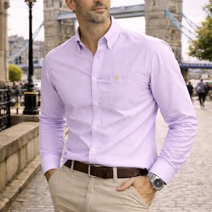 Puede incluir: Camisa abotonada de color lila claro con un peque&ntilde;o emblema amarillo en el bolsillo del pecho. La camisa se combina con pantalones color caqui y un cintur&oacute;n marr&oacute;n. Se ve un reloj plateado en la mu&ntilde;eca.
