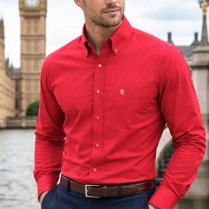 May include: A bright red button-down shirt with a small embroidered emblem on the chest pocket. The shirt is paired with navy blue pants and a brown leather belt. The background features a famous clock tower.