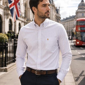 Puede incluir: Una camisa de manga larga con botones, un patr&oacute;n de rayas verticales blancas y azul claro, cuello abotonado y un bolsillo en el pecho con un peque&ntilde;o emblema bordado en dorado. La camisa est&aacute; metida por dentro de un pantal&oacute;n oscuro con un cintur&oacute;n de cuero marr&oacute;n.
