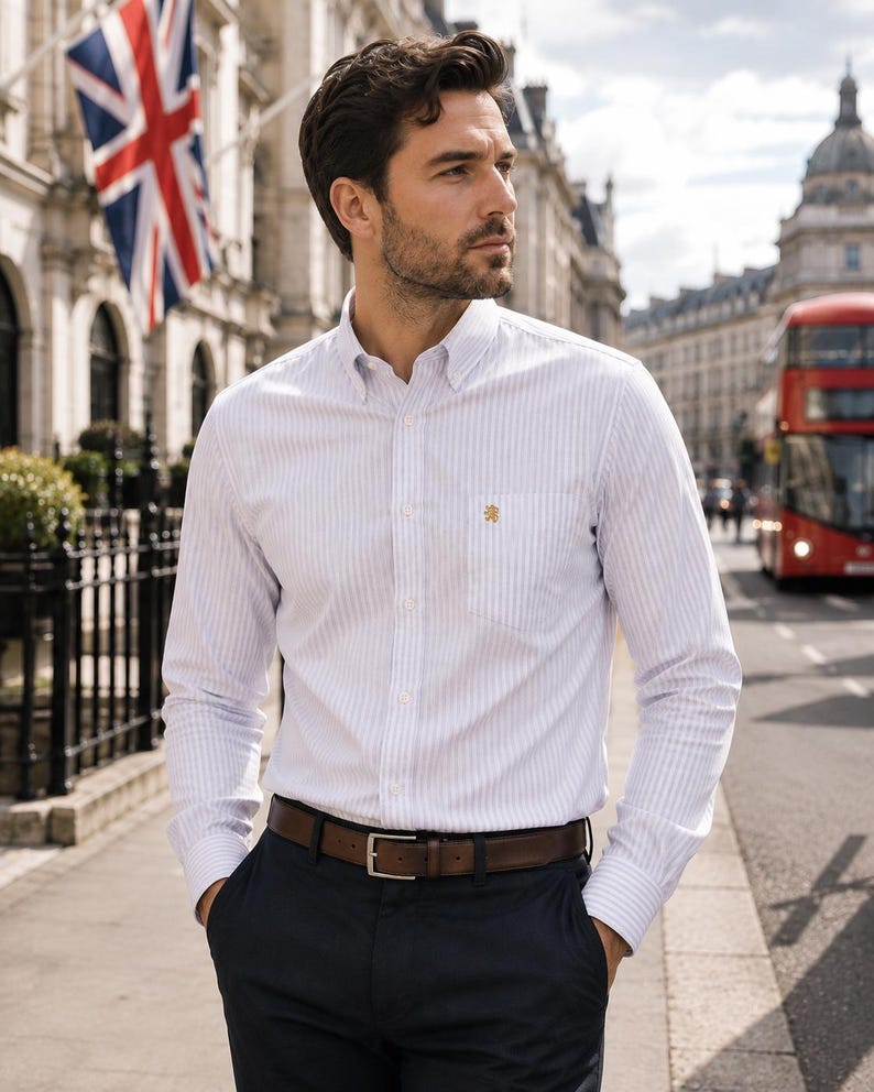 Puede incluir: Una camisa de manga larga con botones, un patr&oacute;n de rayas verticales blancas y azul claro, cuello abotonado y un bolsillo en el pecho con un peque&ntilde;o emblema bordado en dorado. La camisa est&aacute; metida por dentro de un pantal&oacute;n oscuro con un cintur&oacute;n de cuero marr&oacute;n.