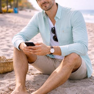 May include: A man wearing a light blue button-down shirt, white t-shirt, and khaki shorts sits on a sandy beach. He holds a black smartphone and wears a brown leather watch with a white face. Sunglasses are tucked into his shirt.