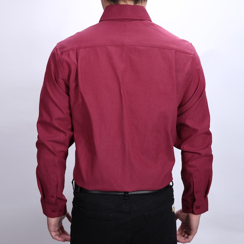 May include: A burgundy long-sleeved shirt with a button-down collar. The shirt is made of a textured fabric and has buttoned cuffs. The person is wearing black trousers and a black belt.