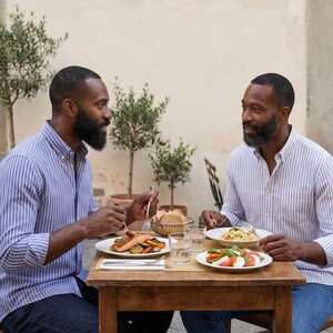 Op de afbeelding: Twee personen dineren in de open lucht aan een houten tafel. De maaltijd omvat zalm, groenten, pasta en een caprese salade. Ze dragen blauw-wit gestreepte shirts. Brood en waterglazen staan ook op tafel.