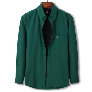 Peut inclure: Une chemise boutonn&eacute;e &agrave; manches longues vert &eacute;meraude fonc&eacute;. La chemise pr&eacute;sente un col boutonn&eacute;, une poche poitrine et un petit embl&egrave;me brod&eacute; sur la poche. La chemise est suspendue &agrave; un cintre en bois.