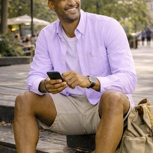 Puede incluir: Una persona con una camisa abotonada de color p&uacute;rpura claro, una camiseta blanca y pantalones cortos color caqui, sosteniendo un tel&eacute;fono inteligente negro. Lleva un reloj con una correa negra y una pulsera plateada. Tambi&eacute;n se ve una mochila color canela y zapatillas blancas.