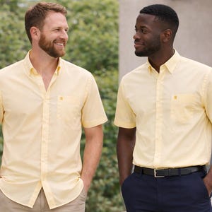 Puede incluir: Dos hombres con camisas amarillas claras de manga corta con botones. Las camisas tienen un bolsillo en el pecho y se combinan con pantalones caqui y azul marino. Están de pie al aire libre frente a un fondo verde frondoso.