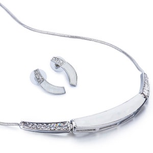 Puede incluir: Un collar de plata con un colgante rectangular blanco curvo y una fila de cristales transparentes. El collar viene con un par de pendientes a juego con un diseño rectangular blanco curvo y cristales transparentes.