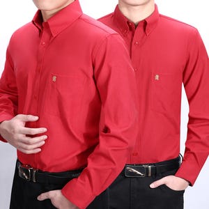 Könnte beinhalten: Zwei langärmelige rote Hemden mit einem kleinen goldenen Logo auf der linken Brusttasche. Die Hemden haben einen Knopfkragenshirt und Knopfmanschetten.