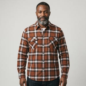 May include: A long-sleeve button-down shirt featuring a brown, black, and white plaid pattern. The shirt includes a standard collar, a front button placket, and two chest pockets with buttoned flaps. The sleeves have buttoned cuffs and roll-up tabs.