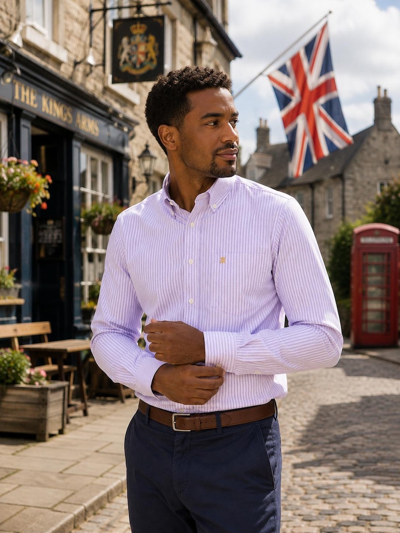 Puede incluir: Una camisa de manga larga con botones, un patr&oacute;n de rayas verticales en color morado claro y blanco, cuello abotonado y un bolsillo en el pecho con un peque&ntilde;o logotipo bordado. La camisa est&aacute; metida por dentro de un pantal&oacute;n azul oscuro con cintur&oacute;n de cuero marr&oacute;n.