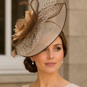 May include: A wide-brimmed fascinator hat in shades of brown and beige, featuring a structured circular base with a dark brown trim. The hat is adorned with a layered arrangement of light brown feathers, sheer polka-dot netting, and loops.