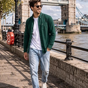 Peut inclure: Une personne v&ecirc;tue d'une chemise vert fonc&eacute;, d'un t-shirt blanc, d'un jean bleu clair et de baskets blanches. Le Tower Bridge de Londres est visible en arri&egrave;re-plan par une journ&eacute;e ensoleill&eacute;e.
