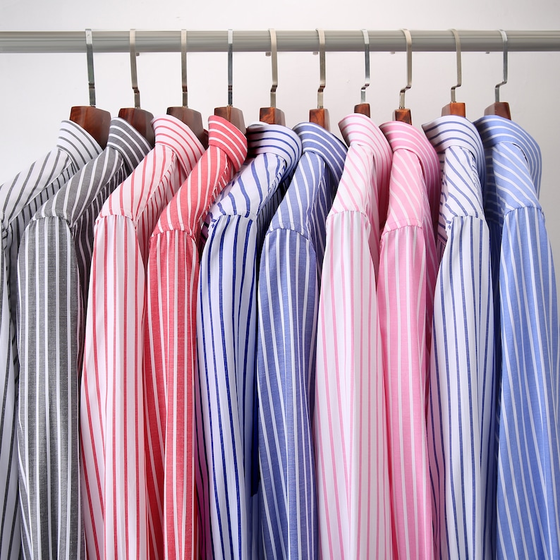 Op de afbeelding: Een verzameling gestreepte overhemden in verschillende kleuren, waaronder grijs, rood, blauw en roze, hangend aan een metalen stang met houten kleerhangers. De overhemden hebben verticale strepen van verschillende breedtes.
