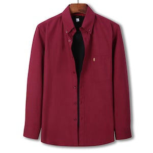 May include: A long-sleeved, button-down shirt in a deep burgundy hue. The shirt has a button-down collar, a chest pocket with a small gold emblem, and a classic fit. It is displayed on a wooden hanger.