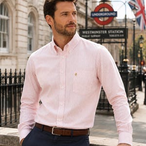 Puede incluir: Una camisa de manga larga con botones, con un patr&oacute;n de rayas verticales en color rosa claro y blanco, cuello abotonado y un bolsillo en el pecho con un peque&ntilde;o emblema bordado en dorado. La camisa est&aacute; metida en un pantal&oacute;n azul marino oscuro con un cintur&oacute;n marr&oacute;n.