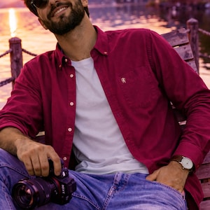May include: A man in a burgundy shirt, white t-shirt, and blue jeans, wearing sunglasses and a wristwatch. He is holding a camera. The background features a lake and mountains, with a sunset in the distance.