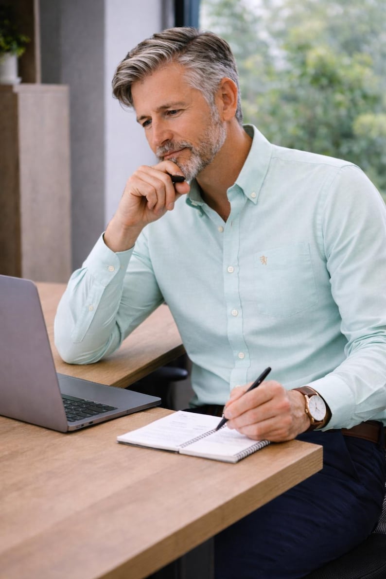 May include: A light green button-down shirt with a small embroidered emblem on the chest pocket. The shirt is worn with a brown leather watch and a notebook. A laptop is on the desk.