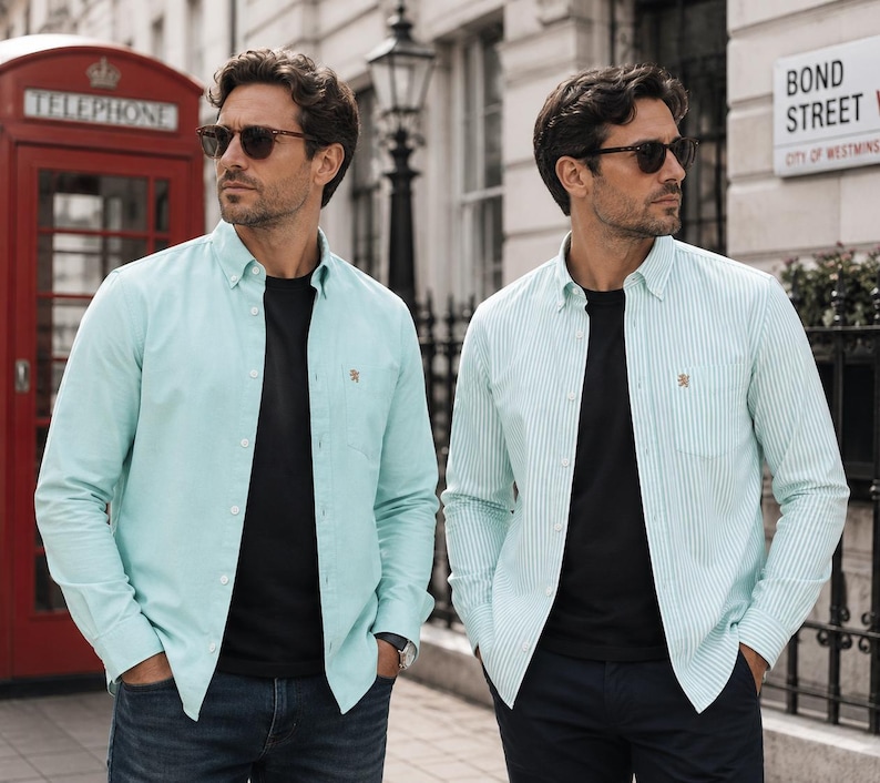Puede incluir: Dos camisas de manga larga con botones sobre camisetas negras. Una camisa es de color verde menta claro liso y la otra presenta un patr&oacute;n de rayas verticales en verde menta claro y blanco. Ambas tienen un peque&ntilde;o le&oacute;n dorado bordado en el pecho.