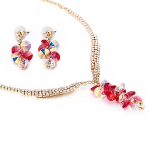 May include: A gold-toned necklace and earring set. The necklace and earrings feature clusters of red, pink, and iridescent crystals. The necklace has a delicate chain with embedded clear crystals. The earrings are post-style.