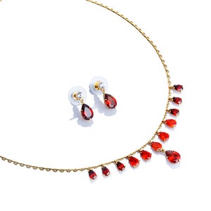 May include: A gold necklace with a delicate chain and red teardrop-shaped gemstones. Matching earrings with red teardrop-shaped gemstones and a single clear gemstone.