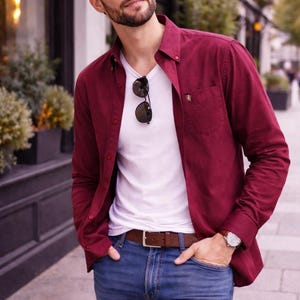 May include: A man in a burgundy shirt over a white V-neck, with blue jeans and a brown leather belt. Sunglasses are on the neckline. A wristwatch is visible. The outfit is suitable for casual wear.