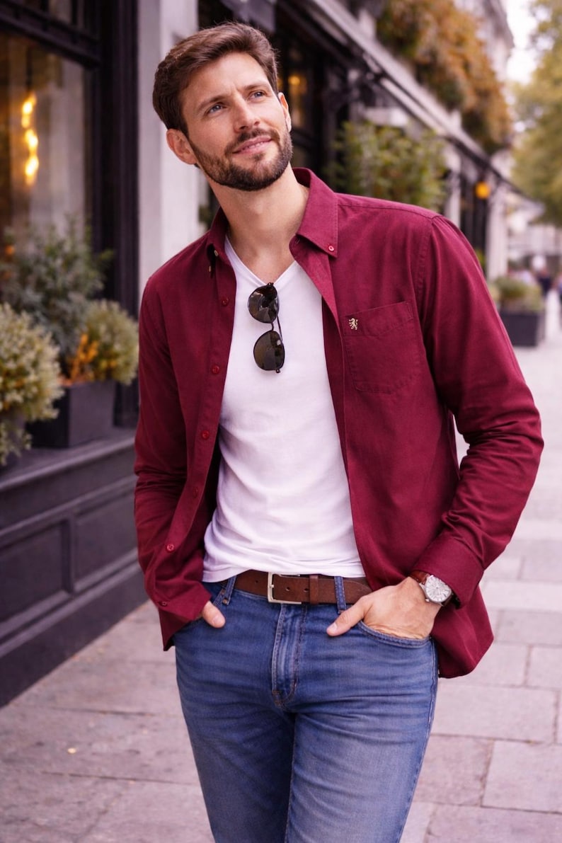 May include: A man in a burgundy shirt over a white V-neck, with blue jeans and a brown leather belt. Sunglasses are on the neckline. A wristwatch is visible. The outfit is suitable for casual wear.