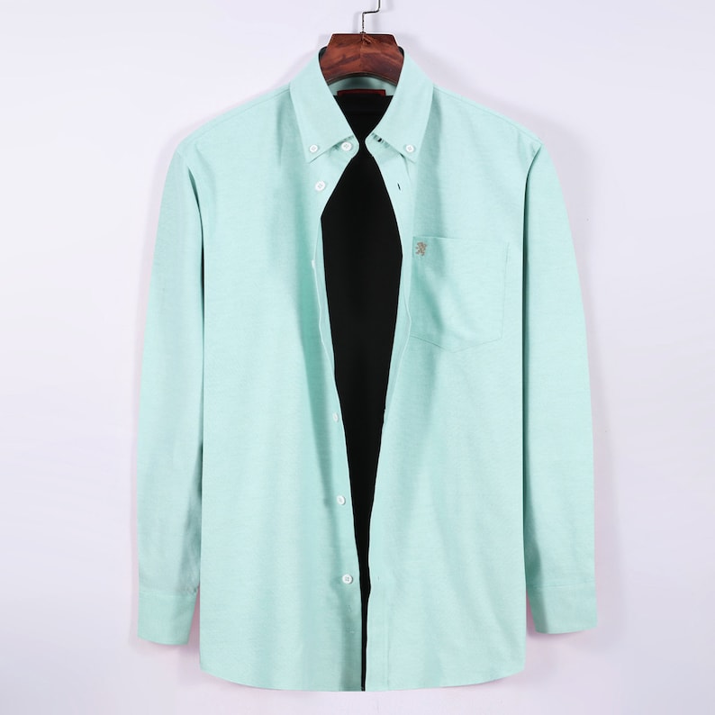 May include: A light green button-down shirt with long sleeves, a chest pocket, and a small embroidered emblem. The shirt is hanging on a wooden hanger against a white background. The shirt has a classic collar and buttoned cuffs.