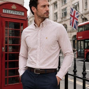 Puede incluir: Una camisa de manga larga con botones y un patr&oacute;n de rayas verticales en blanco y beige, con cuello abotonado, un bolsillo en el pecho con un peque&ntilde;o logotipo bordado y una tapeta frontal, combinada con pantalones azul marino oscuro.