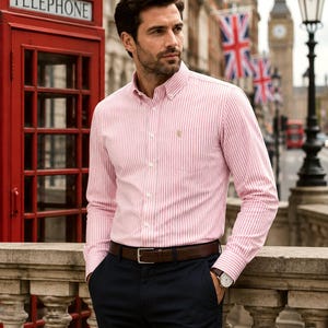 Puede incluir: Una camisa de manga larga con botones, un patr&oacute;n de rayas verticales rosas y blancas, cuello abotonado y un peque&ntilde;o logotipo bordado en el bolsillo del pecho. La camisa est&aacute; metida en un pantal&oacute;n azul marino oscuro con un cintur&oacute;n de cuero marr&oacute;n.