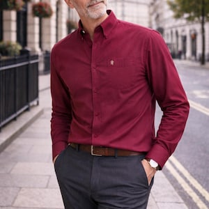May include: A deep red long-sleeved shirt with a button-down collar and a small emblem on the chest pocket. The shirt is worn with grey trousers and a brown leather belt. A wristwatch is visible. The outfit is suitable for business or casual wear.