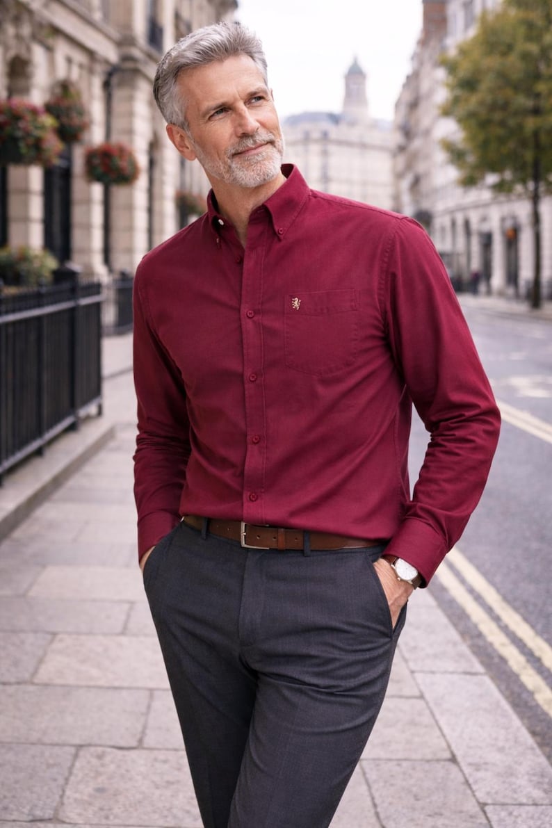 May include: A deep red long-sleeved shirt with a button-down collar and a small emblem on the chest pocket. The shirt is worn with grey trousers and a brown leather belt. A wristwatch is visible. The outfit is suitable for business or casual wear.