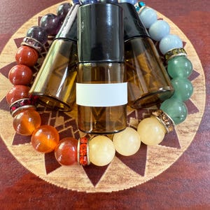 Puede incluir: Botellas de rodillo de vidrio ámbar con tapas negras sobre un posavasos de madera, rodeadas por una pulsera de chakras. La pulsera presenta una variedad de cuentas de piedras preciosas de colores, incluyendo rojo, naranja, amarillo, verde, azul y morado.