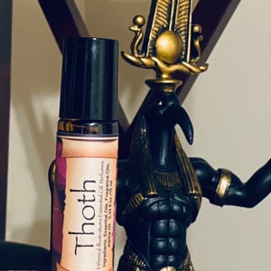 May include: A small brown glass bottle with a black rollerball top. The bottle is labeled "Thoth" and contains essential oils. The bottle is sitting on a wooden surface in front of a gold and black statue.