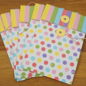May include: A set of six colorful paper gift bags with polka dots and stripes. The bags are decorated with pink, yellow, green, and blue accents. Each bag has a small button closure.