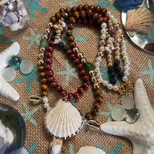 Puede incluir: Un collar de múltiples hilos con cuentas marrones, rojas y blancas, detalles dorados y un gran colgante de concha blanca. El collar se muestra sobre un fondo de arpillera con conchas y estrellas de mar.