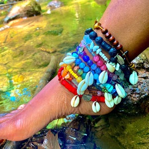 May include: A colorful ankle bracelet with multiple strands of beads, shells, and charms. The bracelet features a rainbow of colors, including red, orange, yellow, green, blue, and purple. The bracelet is adorned with small white shells and gold-colored charms.