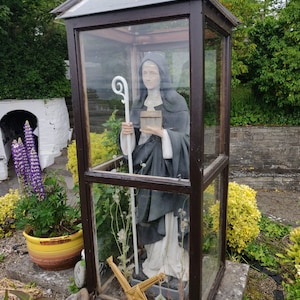 May include: A white statue of a nun holding a book and a staff, inside a glass case with a wooden frame. The statue is surrounded by dried plants and a straw cross.