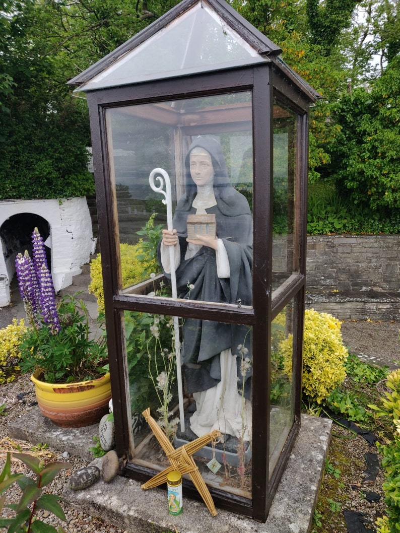 May include: A white statue of a nun holding a book and a staff, inside a glass case with a wooden frame. The statue is surrounded by dried plants and a straw cross.