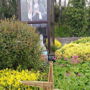 May include: A statue of a nun in a glass enclosure, surrounded by green plants and a woven cross made of natural materials.