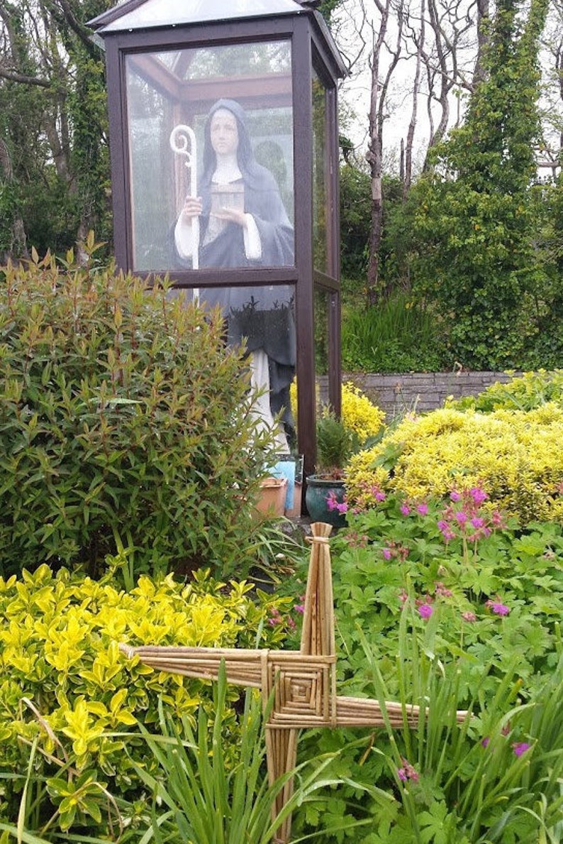 May include: A statue of a nun in a glass enclosure, surrounded by green plants and a woven cross made of natural materials.