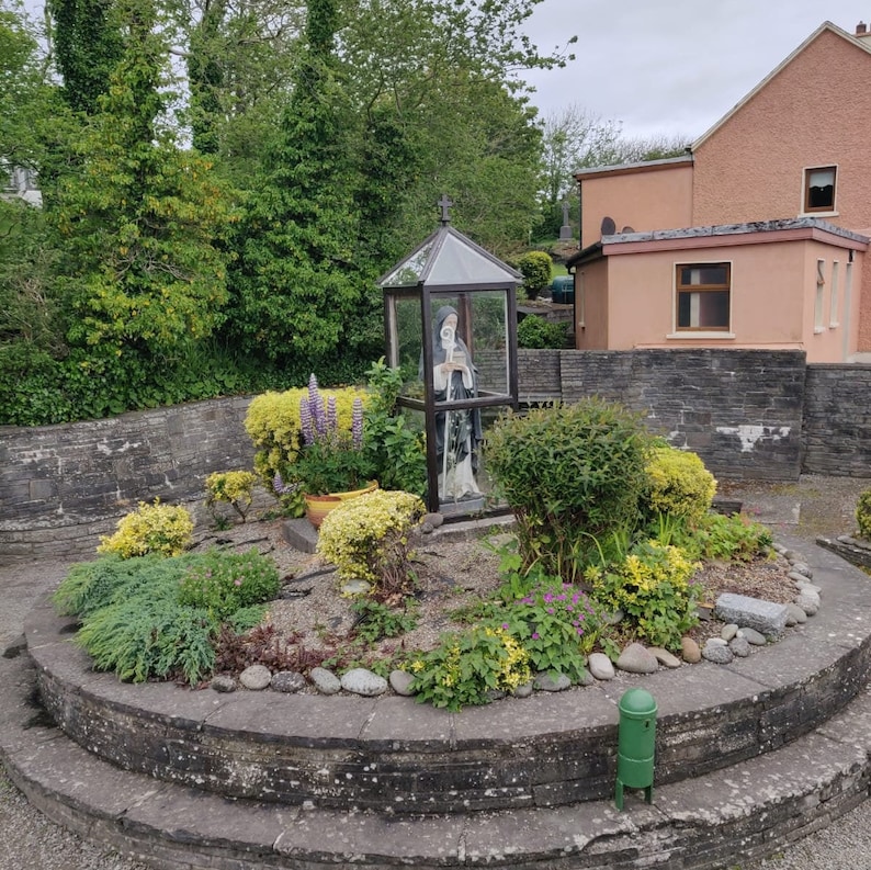 May include: A statue of a woman in a white robe and a blue cloak is enclosed in a glass case. The statue is surrounded by a circular garden with green plants and flowers. The garden is surrounded by a stone wall.