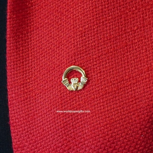Könnte beinhalten: Eine goldfarbene keltische Knoten-Anstecknadel auf rotem Stoffhintergrund. Der Knoten ist ein traditionelles Symbol für Liebe, Freundschaft und Ewigkeit.