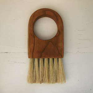Può includere: Una spazzola in legno con un manico con una grande apertura circolare. La testina della spazzola è rettangolare con una serie di setole color naturale. Il legno ha un caldo tono marrone-rossastro.