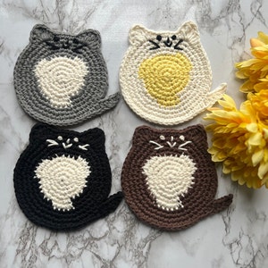 May include: Four crocheted cat-shaped coasters in gray, white, brown, and black. The coasters are made with a circular design and have a white center with a crocheted cat face.