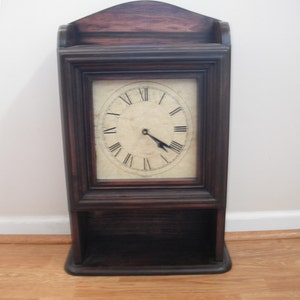 Peut inclure: Une horloge murale en bois brun foncé avec un cadran blanc et des chiffres romains noirs. L'horloge a une étagère en dessous.