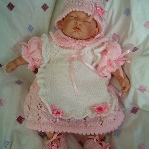 May include: A baby doll wearing a pink knitted outfit, including a hat, dress, and shoes. The dress has a white bodice with a pink apron and is adorned with pink ribbons and small flower accents. The doll is lying on a white surface.