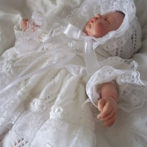 Może przedstawiać: Biała dzianinowa sukienka do chrztu dla dziecka z koronkowym wykończeniem i pasującym kapturem. Kapturek jest zawiązany białą satynową wstążką. Sukienka ma długie rękawy z koronkowymi mankietami. Dziecko leży.