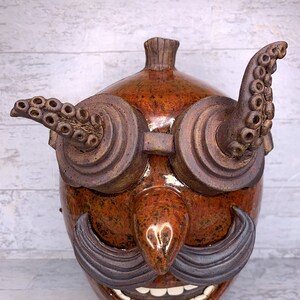 Pode incluir: Uma máscara de cerâmica caprichosa com um acabamento salpicado castanho e laranja. A máscara apresenta óculos grandes e redondos com braços em forma de tentáculos, um nariz proeminente, um bigode cinzento e um sorriso largo e dentuço. Uma peça decorativa única.